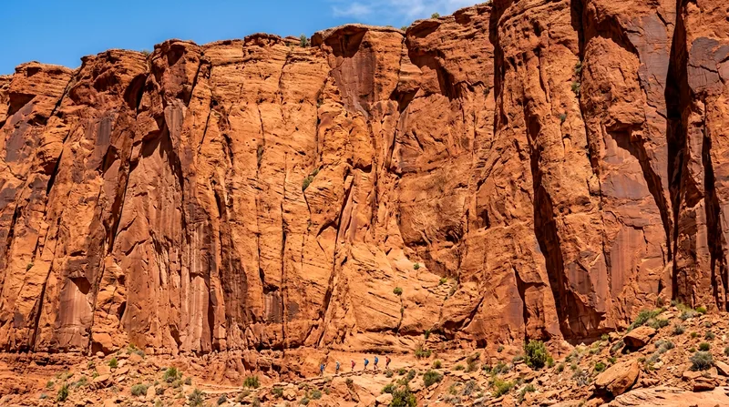Massive red rock cliff face towering above tiny distant hikers on trail below.