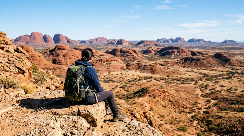 Hiker sitting on rocky outcrop viewing multiple Kata Tjuta domes across landscape.