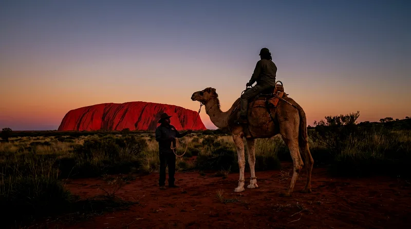 Mounted rider on camel at sunset with Uluru glowing red on horizon and handler standing by.