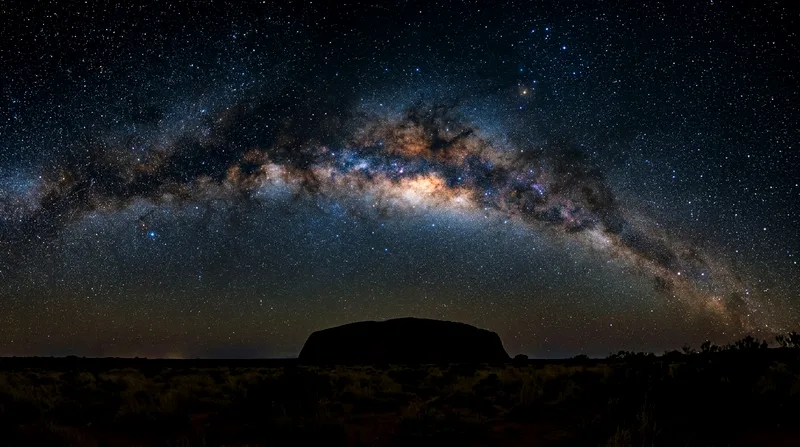 Milky Way arcing across night sky above darkened Uluru silhouette.